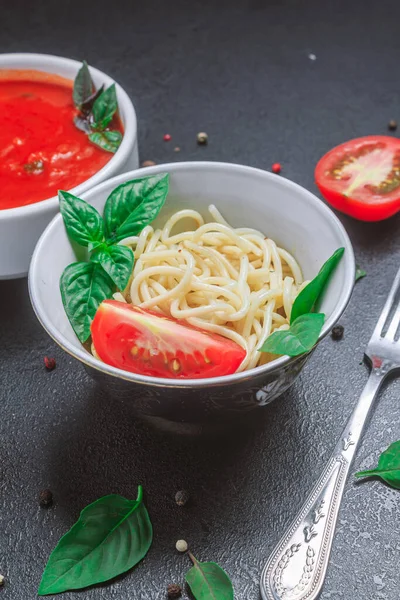 Špagety v talíři s rajčatovou omáčkou v malé misce s kořenící bazalkovou omáčkou na černém stole. kopírovat prostor, zavřít, svislá orientace — Stock fotografie