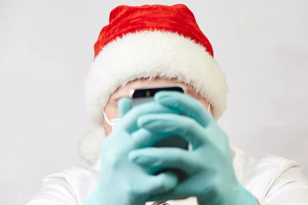 Концепція коронавірусу, людина в капелюсі Санта та медична маска з телефоном на білому тлі — стокове фото