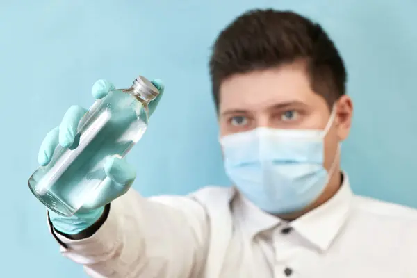 Coronavirus-Konzeptmensch in medizinischer Maske blickt auf Flasche auf blauem Hintergrund — Stockfoto