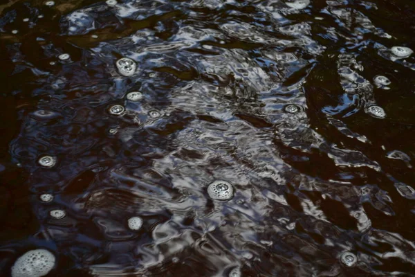 Slightly agitated surface of the river. The river is saturated with iron, the water is brown. White bubbles in the water. Play of colors of white, blue and brown.