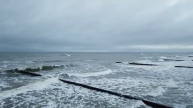 Şiddetli Baltık Denizi kıyısında, bulutlu bir kış gününde, ağır ahşap yığınların üzerinde dalgalar yuvarlanırken bulutlu bir gökyüzü alçalıyordu. İnsansız panoramik çekim olmadan..