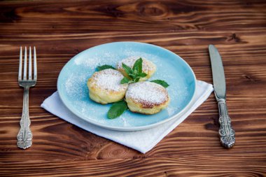 Çatal ve bıçaklı mavi tabakta cheesecake, kahverengi ahşap arka planda pamuk bez.