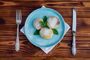 Çatal ve bıçaklı mavi tabakta cheesecake, kahverengi ahşap arka planda pamuk bez.