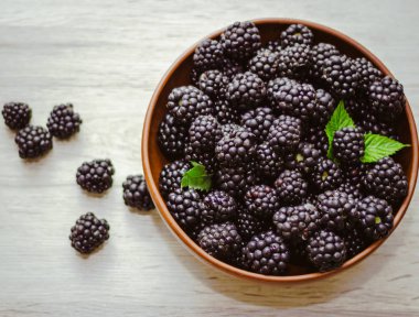 Olgun böğürtlenler, hafif ahşap arka planda kil bir kasede yapraklarla. Düz yerde, üst tarafta. Ahşap masadaki kil kasede böğürtlen fotoğrafı. Yüksek çözünürlüklü ürün.