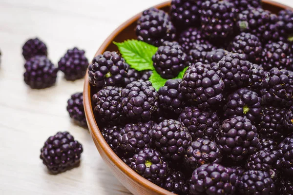 Ripe blackberries with leaves in a clay bowl on a light wooden background. Flat lay, top view. Photo of blackberry in clay bowl on wooden table. High resolution product.