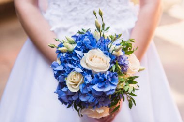 Gelinin elinde düğün buketi, beyaz bir elbisenin önünde.