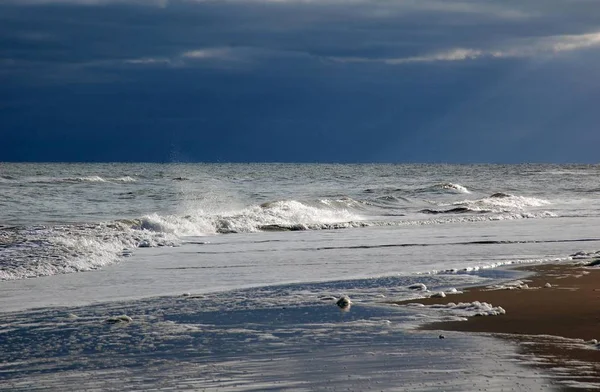Bulutlu gökyüzünün altında kumda kırılan dalgalar.