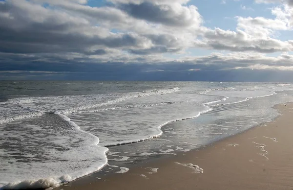 Bulutlu gökyüzünün altında kumda kırılan dalgalar.
