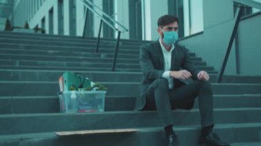 Merdivenlerde oturan maskeli bir adam kişisel eşyalarla dolu bir kutuyla ve üzerinde 
