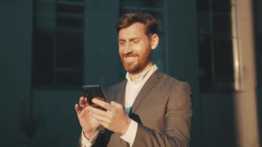 Gün ışığında genç, mutlu adam şehir merkezinde duruyor. Telefon mesajları, kayan gülümseme teknolojisi, güneşli günler, yavaş çekim başarısı kullanıyor.