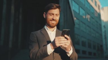Şehir merkezindeki mutlu işadamı duruşu telefon mesajlaşması, kayan gülümseme teknolojisi, güneşli gün başarısı yavaş çekim kullanıyor.