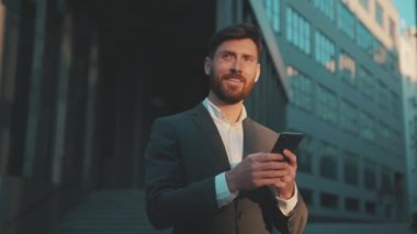 Şehir merkezindeki iş adamı durağında çekim yapmak için telefon mesajlaşması, kaydırma, güler yüzlü gülümseme teknolojisi, güneşli gün başarısı, ağır çekim.
