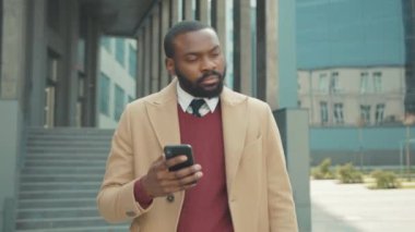 Ciddi Afro-Amerikalı genç çekici adam, gün batımında sokakta yürüyen telefon kullanıyor. Cep telefonu, bilgisayar, internet teknolojisi, iş adamları, siyahi şehir gezginleri...