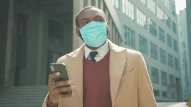 Yüz maskesi takan genç Afrikalı bir adam sokak koronavirüsüne telefon kullanıyor. Güvenlik önlemi, salgın salgını, tıbbi maske, yavaş çekim.