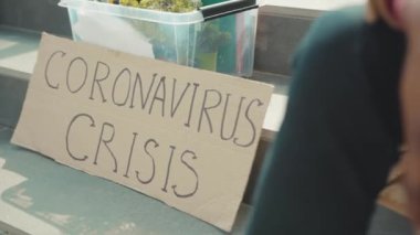 Afrika kökenli Amerikalı genç adam maske takıyor. Elinde kişisel eşyalar ve Coronavirus Krizi yazılı posterle merdivenlerde oturuyor. İşsiz depresyon ekonomisi ağır çekim.