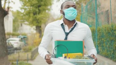 Afrikalı Amerikalı ofis çalışanı maske takıyor. Elinde kişisel eşyalarla yürüyor. Coronavirus krizi yüzünden işten atıldı.