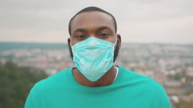 Sağlıklı, koruyucu cerrahi maske takan Afro-Amerikan bir portre şehrin tepesinde tepede duruyor. 2020 Salgını. Coronavirus. Karantina.