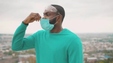 Genç Afro-Amerikan iş adamını uyarın. Koruyucu tıbbi maske takıyor ve şehir manzarasının üzerinde duran gözlük takıyor. Geçmişi. Karantinaya alın. Sağlık hizmetleri.