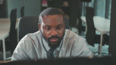 Yakışıklı Afro-Amerikan yakışıklı adam modern ofis programcılarında bilgisayar kullanıyor. Monitör işadamına ciddi ciddi bakıyor. Yavaş çekim.