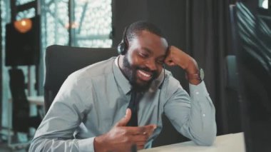 Komik Afro-Amerikalı yakışıklı adam kulaklığı iş için kullanıyor bilgisayarla konuşuyor modern ofis programcısı gülümsüyor ekranda çalışan başarılı iş adamına bakıyor.