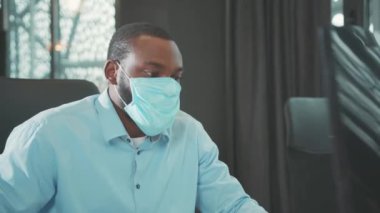 Maske takan ciddi bir Afrikalı Amerikalı modern ofis programcılarında bilgisayar kullanıyor. Monitör iş adamı Coronavirus 'a bakıyor. Yavaş çekim.