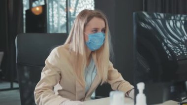 Masaüstü bilgisayarında çalışan, virüs önleme klavyesi için cerrahi maske takan ofis sarışın genç bayan çalışanı. Salgın. Sağlık hizmeti kavramı.