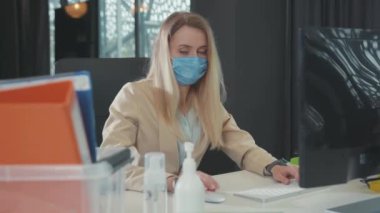 Ofisteki bilgisayarda çalışan yüz maskeli beyaz kadın. Mutsuz sarışın kız masasında umutsuzluk içinde oturarak işini kaybetti. Küresel kriz 2020.