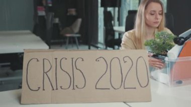 İşten ayrılan ve ofisten kişisel eşya kutusuyla ayrılan genç bir kadın. Kriz 2020 afişi çalışma masasında. Küresel işsizlik. Kilitlenme. Karantina.