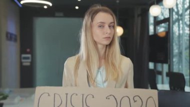 Boş işyerinde Crisis 2020 karton afişini gösteren ciddi işsiz genç bir kadın. Küresel işsizlik. Finansal felaket. Coronavirus karantinası.