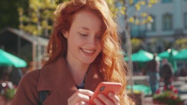 Güzel, genç, kızıl saçlı, beyaz kız öğrenci akıllı telefon sosyal medya uygulaması kullanıyor. Mesajlaşan gülümseme, güneşli bahar gününde dışarıda duruyor. Şehir halkı yaşam tarzı.
