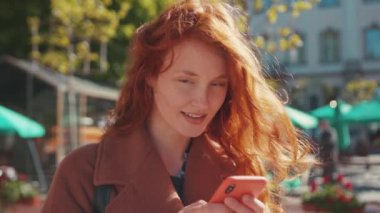 Açık havada çekingen, ceketli, elinde akıllı telefonuyla neşeyle gülümseyen sevimli kızıl kızın portresi. Gençler. Moda. Kentsel yaşam tarzı.