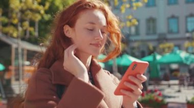 Güzel görünümlü, kızıl saçlı, beyaz bir kadın akıllı telefondan olumlu haber mesajı okuyor. Güzel şehir merkezinde kaldığı için seviniyor. Portreler. Kentsel yaşam tarzı.