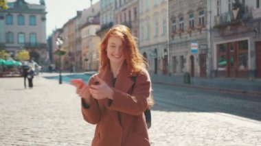 Genç, neşeli kızıl saçlı, palto giyen, dans eden, açık havada akıllı telefon ağı cihazıyla oynayan kız. Eğlenceli bir konsept. Kullanıcı arayüzü ile canlandırma için.