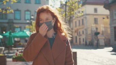Sağlık maskesi takan, kendine güvenen genç, kızıl saçlı kadın şehir merkezinde yürürken akıllı telefon kullanıyor. Covid-19. Salgın.