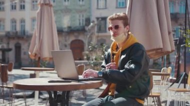 Kafe terasında portatif dizüstü bilgisayarla çalışan yüz maskeli genç beyaz bir adam. Sosyal uzaklık. Bağımsız yaşam tarzı. Coronavirüs.