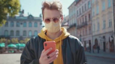 Güneş gözlüğü takan koruyucu maskeli genç turist ya da öğrenci güneşli bir havada şehir merkezindeki sokakta akıllı telefon uygulaması kullanıyor..
