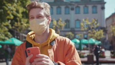 Sarışın, genç ve yakışıklı bir adam, Coronavirus 'a karşı koruyucu bir maske takıyor ve dışarıdaki güzel caddede kalan akıllı telefon zaman çizelgesinde haberler okuyor. Salgın. Sağlık Hizmetleri.