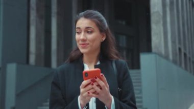 Neşeli, genç, yetişkin, beyaz kadın ofis müdürü modern binanın yanında yürüyor mutlu bir şekilde haberlere bakıyor, akıllı telefon sosyal medyası açık havada..