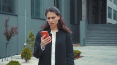 Kafası karışmış beyaz bir kadın akıllı telefondan haber zaman çizelgesi okuyor. Şaşırmış bir yüz ifadesiyle ofis binasının önünde durmakta tereddüt ediyor..