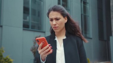 Kafası karışmış beyaz bir kadın akıllı telefondan olumsuz geri bildirim okuyor. Kötü sürpriz mesaj yazma konusunda hayal kırıklığına uğruyor. Açık havada portre.