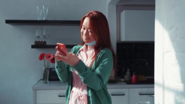 Neşeli Çinli, genç, kızıl saçlı, sıradan kıyafetler giyen, sosyal medyada akıllı telefon ağı kuran, mutfakta duran güzel bir kadın. Sosyal mesafe. Evde kal..