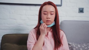 Çekici Asyalı kızıl saçlı sıradan kadın cerrahi yüz maskesi takar ve evde taşınabilir bilgisayarla çalışmaya başlar. Serbest çalışıyorum. Sosyal uzaklık. Sağlık hizmetleri. Koruma.