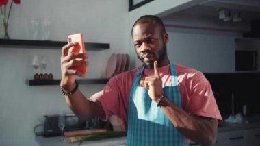 Önlüklü siyahi, çekici bir genç mutfakta akıllı telefondan video görüşmesi yapıyor. Aşçı şefi. Teknoloji ve insanlar. İletişim konsepti.