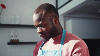 Aperatif giymiş Afro-Amerikan bir adam akıllı telefondan zaman çizelgesi okuyor ve kötü haber içeriğiyle hayal kırıklığına uğruyor. Mutfaktaki adam. Hayal kırıklığı. Teknoloji.