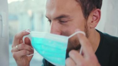 Sıhhiye maskesi takan adam evde oturup karantina koronavirüsündeki sokağa bakıyor kendini tecrit eden depresyon penceresini yavaş çekimde sıkıyor.