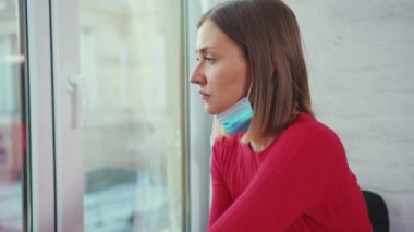 Üzgün genç bir kadın pencere pervazında oturuyor. Tıbbi yüz maskesi takıyor. Koronavirüs kendini tecrit depresyon penceresi.