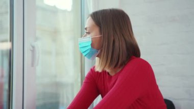 Kapalı bir pencere kenarında oturan genç kadın tıbbi yüz maskesi takıyor. Koronavirüs kendini tecrit depresyon penceresi sıkıcı yavaş çekim.