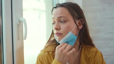 Ciddi genç bir kadın pencere pervazında oturuyor. Tıbbi maske takıyor. Kendini tecrit depresyon penceresi. Sıkıcı yalnızlık, üzücü solunum koronavirüsü.