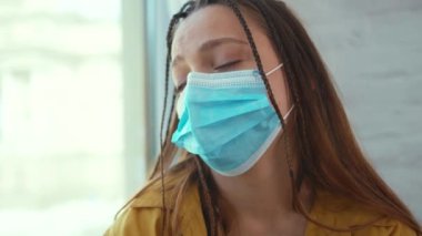 Suratı asık genç bir kadın pencere pervazında oturur tıbbi yüz maskesi takar kendini tecrit depresyon penceresi sıkıcı yalnızlık koruması üzgün solunum koronavirüsü kovid 19 yavaş çekim