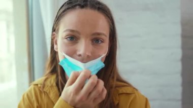 Yakın plan genç kadın Coronavirüs salgınında yüzüne tıbbi maske takıyor. Kamera covid-19 karantina hastalığı salgınına bakıyor.